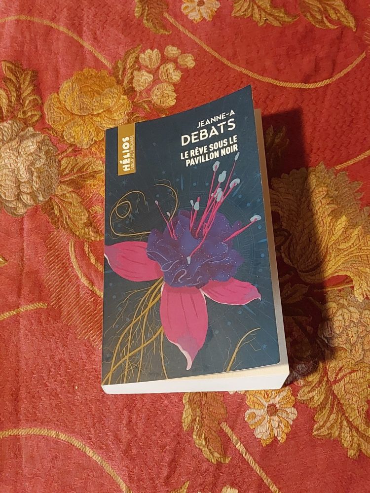 Le livre intitulé Le Rêve sous le Pavillon Noir de Jeanne-A Debats, publié dans la collection Hélios, est posé sur un tissu nacarat (si si) orné de motifs floraux dorés. La couverture du livre représente une fleur de fuchsia stylisée tau cœur violet d’où s’échappent des étamines roses et bleues, sur un fond bleu étoilé.
