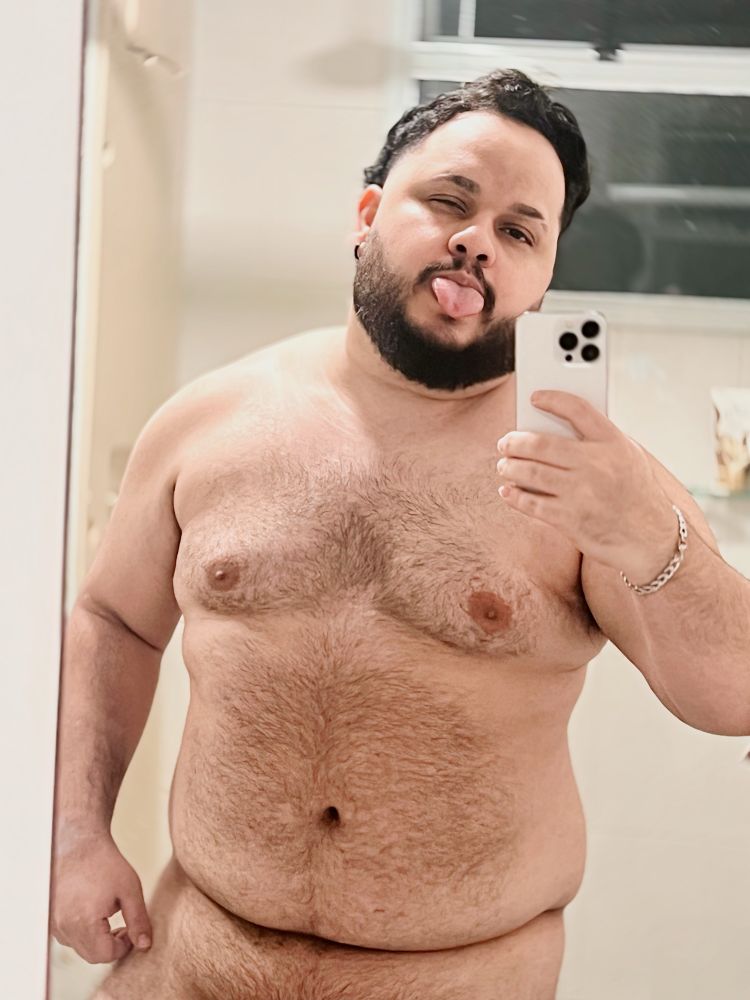 Selfie no espelho, entrando no banho. Estou segurando o celular com a mão direita, olhando pra câmera e com a língua de fora. Cabelo bagunçado. Estou nu e a foto mostra da virilha pra cima.