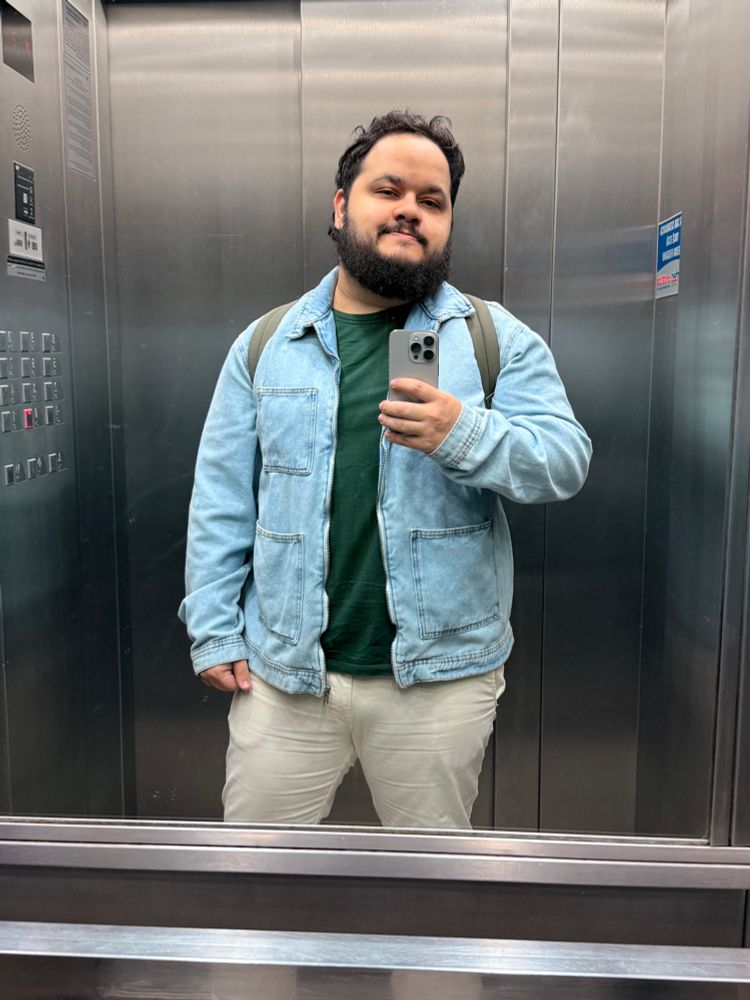 Selfie no elevador. Homem com o cabelo levemente bagunçado, com barba em tamanho médio. Vestindo uma calça bege, camiseta verde escura e jaqueta jeans azul clara. É possível ver alças de uma mochila verde oliva nos ombros.