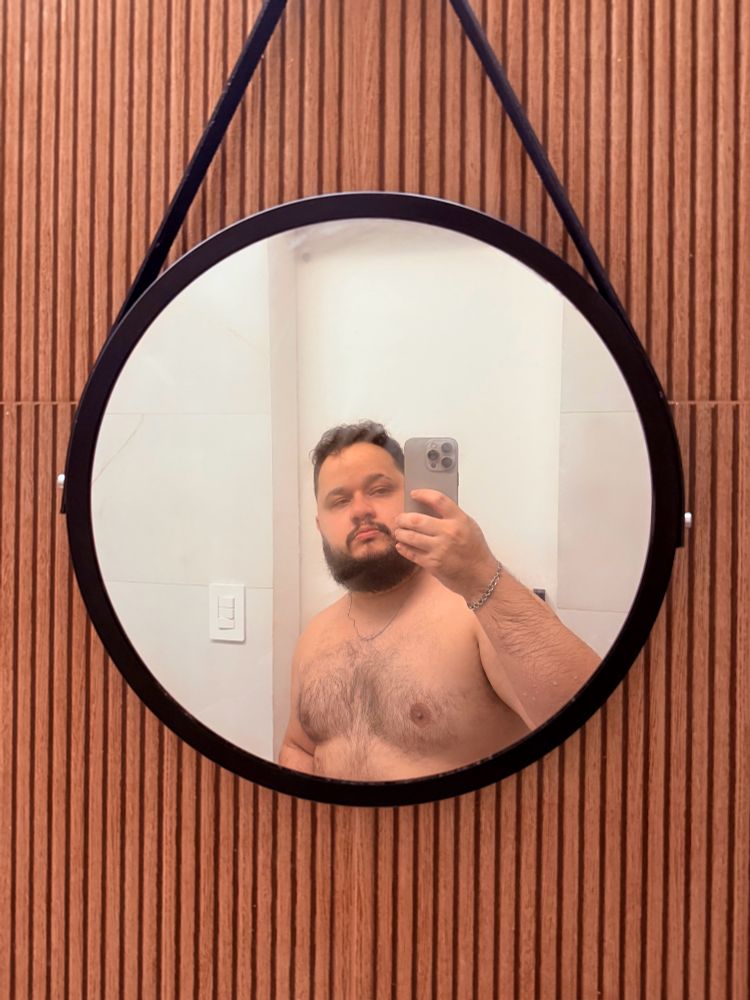 Selfie minha em frente a um espelho redondo de banheiro. O espelho é redondo, com bordas pretas e preso por uma tira de couro preta pendurado na parede. A parede tem um piso marrom que imita madeira estilo ripado. Estou sem camiseta, pós banho, espelho ligeiramente embasado, eu apareço da cintura para cima, com cabelo, barba e pelos do peito ainda molhados. Seguro o celular com a mão direita e aponto para o espelho.