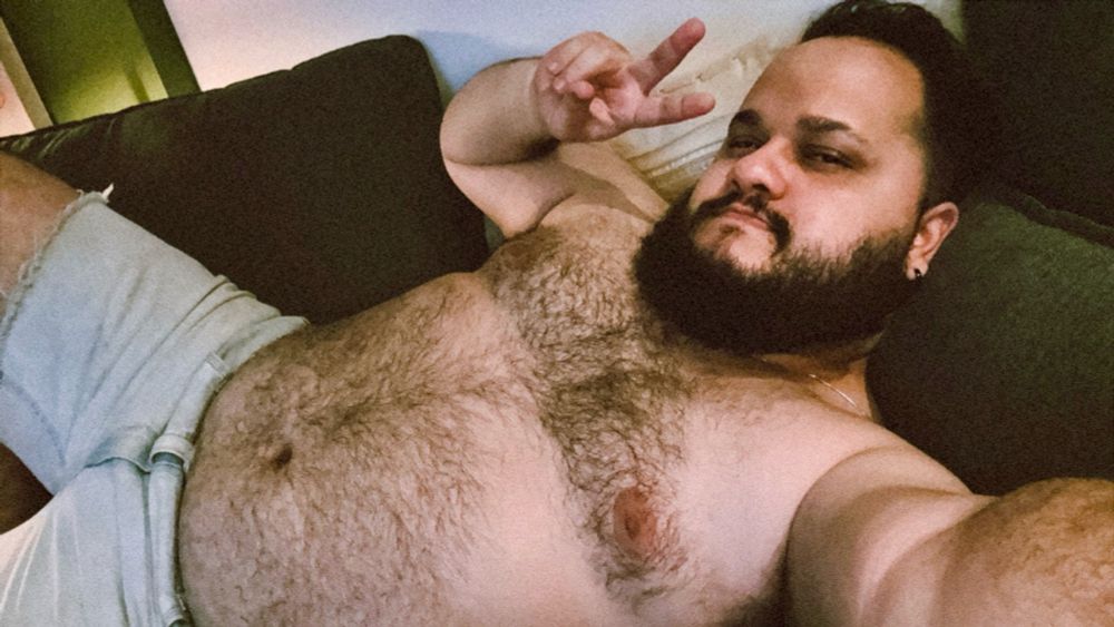 Homem deitado em um sofá escuro, usando shorts jeans claros e sem camisa, exibindo pelos no peito e uma barba cheia. Ele faz um sinal de paz com a mão direita e olha diretamente para a câmera, com expressão descontraída. O ambiente tem iluminação suave.