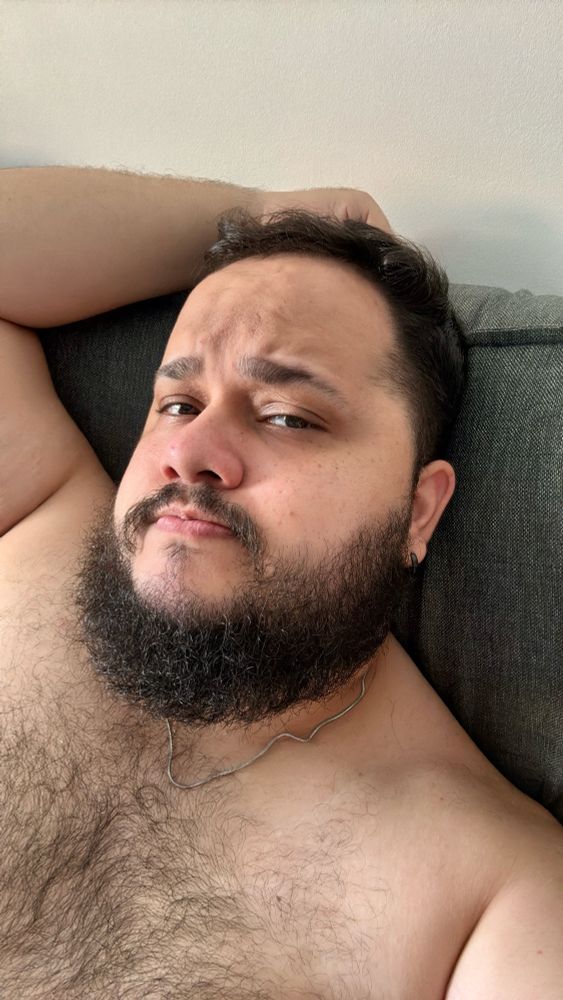 Homem deitado em um sofá cinza, com o braço apoiado atrás da cabeça. Ele está sem camisa, exibindo pelos no peito, barba cheia e expressão levemente pensativa ou desconfiada. A luz natural ilumina o rosto, destacando os detalhes.