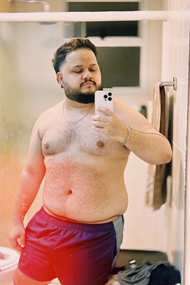 Selfie minha no espelho do banheiro com o box ao fundo, uma toalha pendurada no lado direito e algumas roupas visíveis acima do roupeiro no canto inferior direito. Estou apenas de short na cor verde petróleo, cordão e pulseiras de aço, brincos na cor preta. Estou olhando para o celular enquanto tiro a foto, meu braço direito está erguido (invertido por conta do espelho). Sou baixo e tenho o corpo gordo, ou, como gosta de chamar minha balança, grosso-conjunto — ela diz que eu tenho igual proporção de gordura e músculos, ou algo do tipo, haha. Meu cabelo está cortado como um mullet com a parte de cima e de trás maiores e as laterais menores, sumindo para máquina zero. Tenho barba média e bigode um pouco destacado da barba, pois não encaixa. Estou com os pelos corporais aparados.