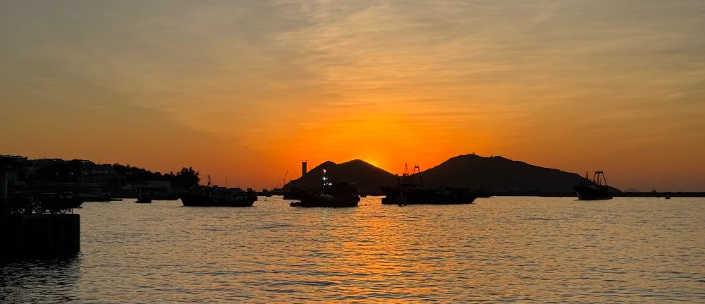 Orange sunset over the ocean with the sun setting behind an island, a few fishing boats in shadow in front.