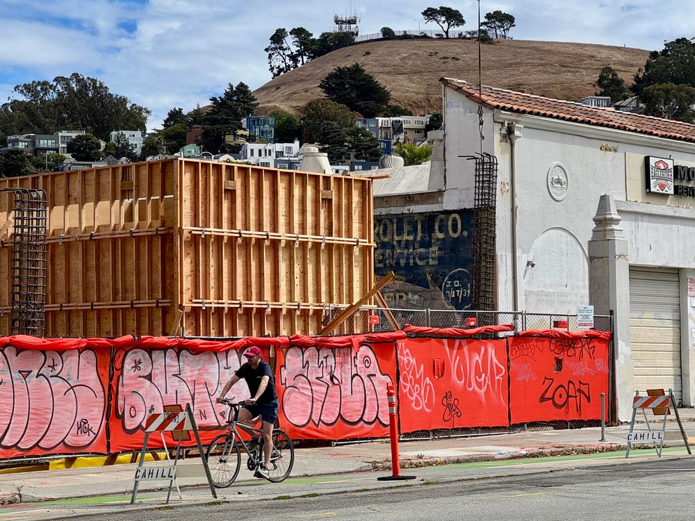 A wider shot of the construction zone. In the photo you can see wooden frames being built for the new building, a crane lowering rebar, the side of the motorcycle shop on Valencia, a cyclist in the bike lane, and Bernal Hill in the background. 