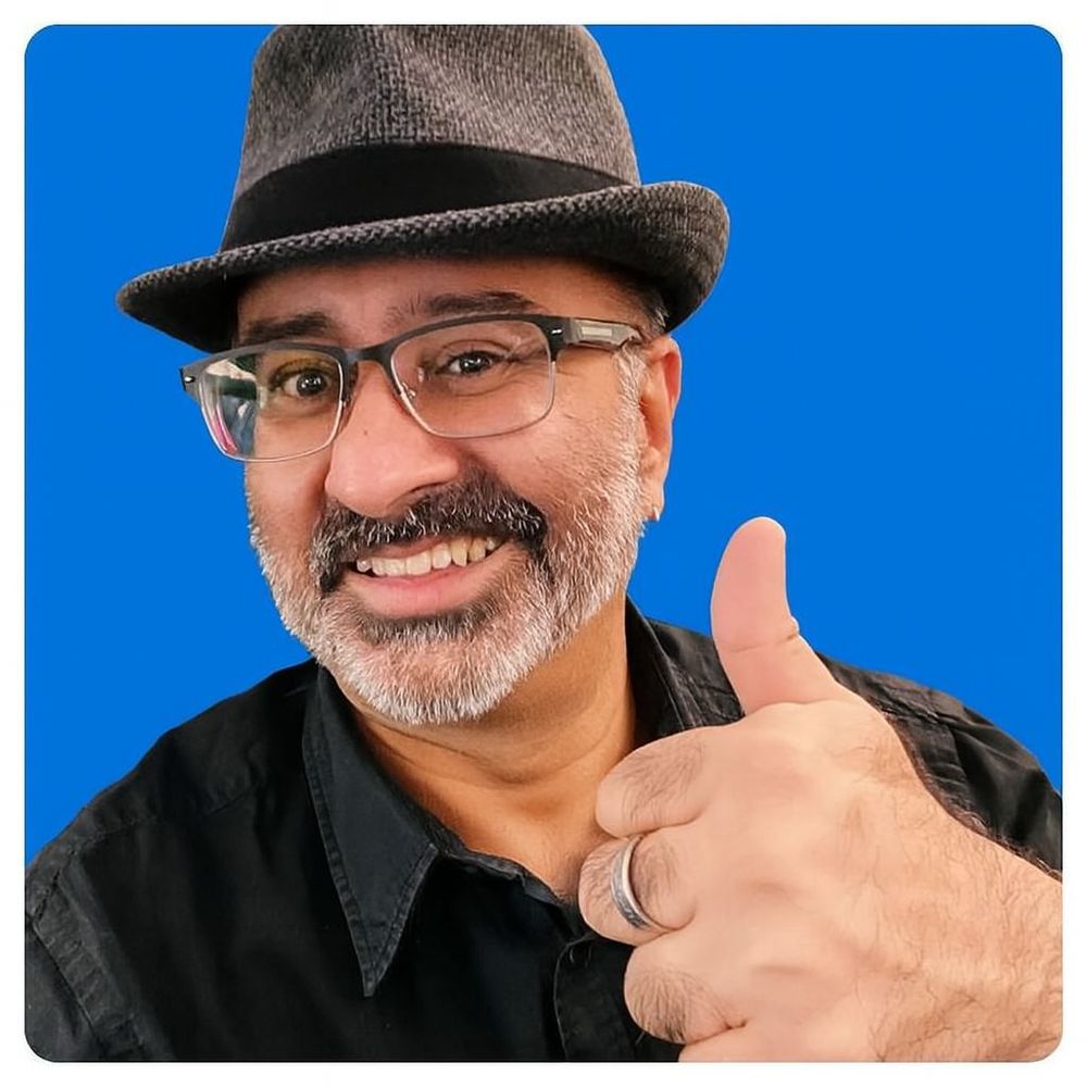 Smiling middle-aged man with a salt-and-pepper beard and glasses giving a thumbs-up gesture against a solid blue background. He is wearing a dark gray fedora hat and a black button-up shirt, and a silver wedding band is visible on his thumb-up hand. The man appears friendly and confident, conveying positivity and approval. Ideal image for themes like encouragement, support, approval, confidence, and personal branding.