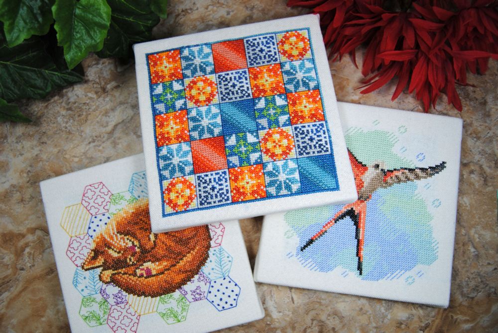 Three square cross stitch designs on stretched canvas, featuring designs inspire by board game covers. Specifically the games Calico, Wingspan, and Azul.