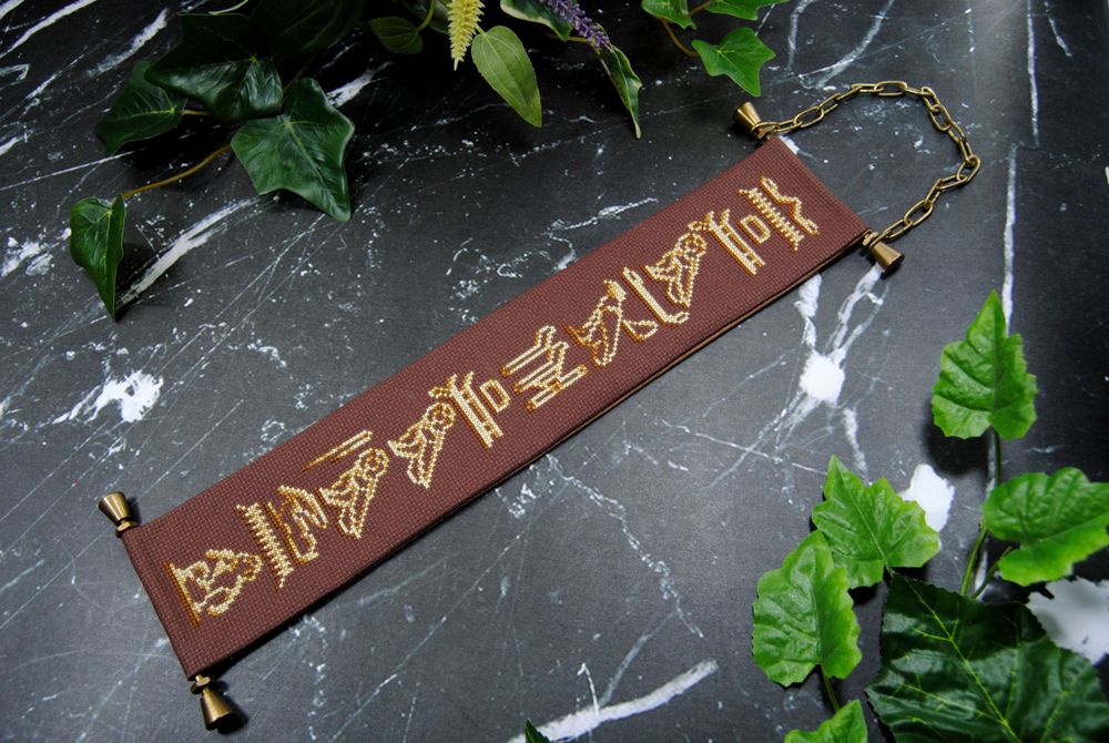 A cross stitch bell pull on dark brown fabric with gold hardware and featuring hieroglyphics from the game Assassin's Creed Origins. In this picture I've attempted to capture the full thing by photographing it diagonally across the frame.