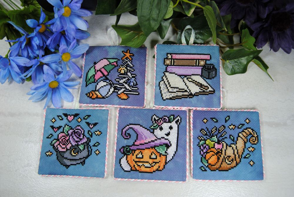 A set of 5 pastel cross stitch ornaments all featuring a similar color palette.  Varying designs, including a summery christmas design with a driftwood tree,  a stack of books with an open journal for writing in.  A cauldron full of flowers, a pumpkin wearing a witch hat hanging out with a catlike ghost, and a cornucopia design.