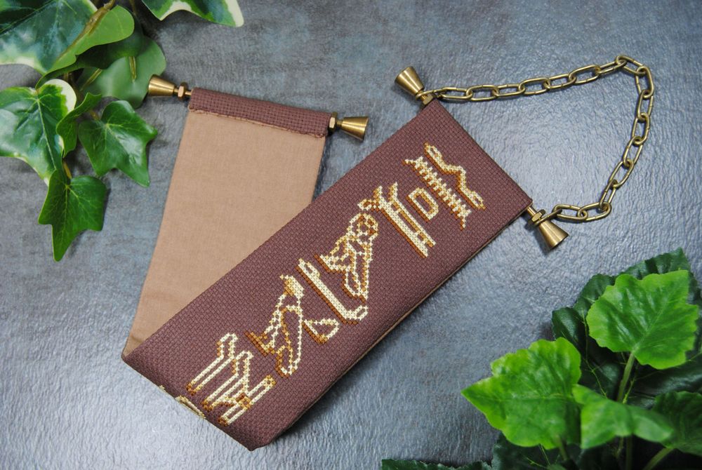 A cross stitch bell pull on dark brown fabric with gold hardware and featuring hieroglyphics from the game Assassin's Creed Origins. In this picture it's folded in half so you can see the back is plain light brown coordinating fabric.