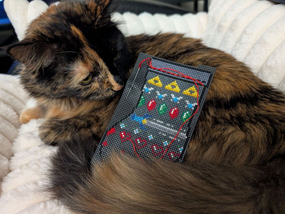 A cross stitch wip of a Zelda themed christmas stocking, propped up against a very curious cat.