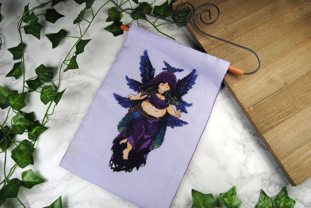 A cross stitched wall hanging on light purple linen featuring a 6-winged Starling fairy designed in collaboration with the artist Frenone.