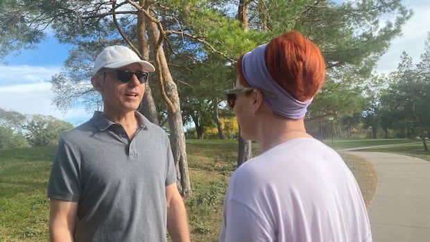 A man in a grey shirt, ball cap and sunglasses can be seen with a woman who has her back turned to the camera.