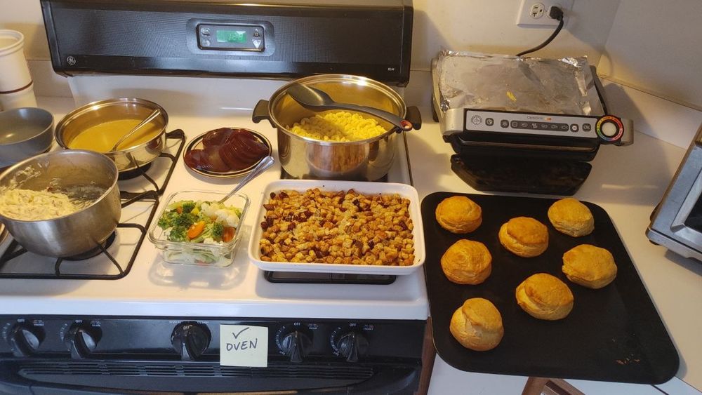 A selection of pots, pans, and trays filled with different helpings of thanksgiving staple foods: from left to right - gravy, mashed potatoes, jellied cranberry sauce, mixed vegetables, macaroni & cheese, stuffing, and biscuits.
