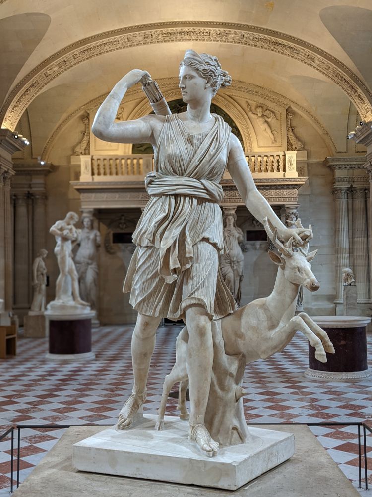 White marble Roman over life sized statue based on an original Greek of the Goddess Artemis with a deer. (The statue is named Artemis, Goddess of the Hunt / Diana of Versailles)
The statue is from circa 325 BCE, and a partial restored Roman copy from the 1st or 2nd century BCE based on a lost Greek bronze original attributed to Leochares.
It is currently displayed in the Louvre museum in Paris.
The statue features the Goddess in a walking movement, hunting near a tree, taking an arrow from her quiver. Her sacred animal, a deer, is jumping next to her. She is wearing a Dorian chiton, sandals and a himation around her waist.
The goddess Artemis is the goddess of hunting, the moon, the wild life, eternal maidenhood and the midwives. She is the daughter of Zeus and the titan Leto, first born of the twin siblings, and helped her mother give birth to her twin brother, Apollo. She is one of Zeus’ favorite daughters with Athena.