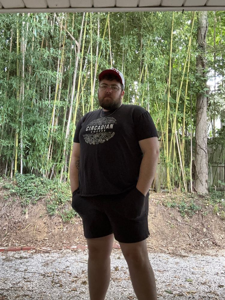 Andy standing in front of bamboo, wearing short black shorts, a black t-shirt with “Circadian Coffee” screenprinted in white, and red and white Cubs hat