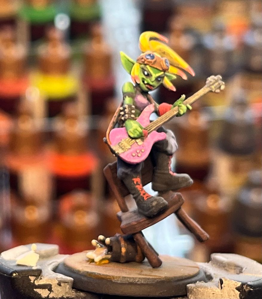 Painted mini of a goblin alchemist ready kin playing a bass and balancing on a chair. 