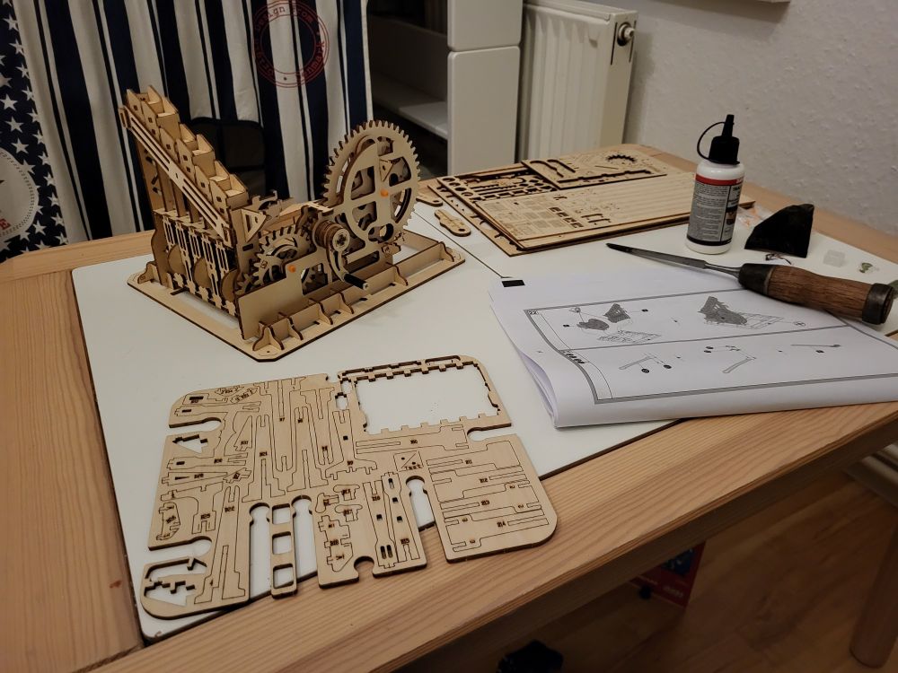 Kleiner Kindertisch mit einem angefangenen 3D-Holzkugelbahn.
Ein Teil des Modell ist schon fertig gebaut mit Zahnräder und Mechanik.
Im Vordergrund ist eine dünne Holzplatte mit gläsernen Teilen. Rechts davon eine Bauanleitung mit einem Stecheisen darauf und dahinter weiter Holzplatten und Holzleim