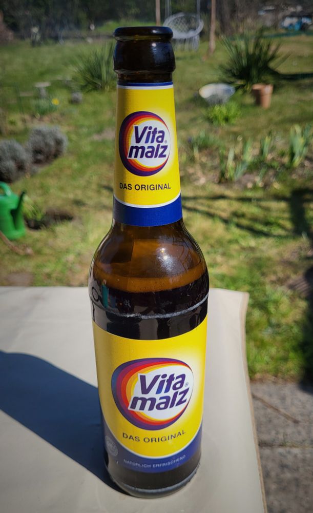 Flasche Vitamalz auf einem Tisch. Im Hintergrund eine Wiese und anderen diversen Gestrüpp 
