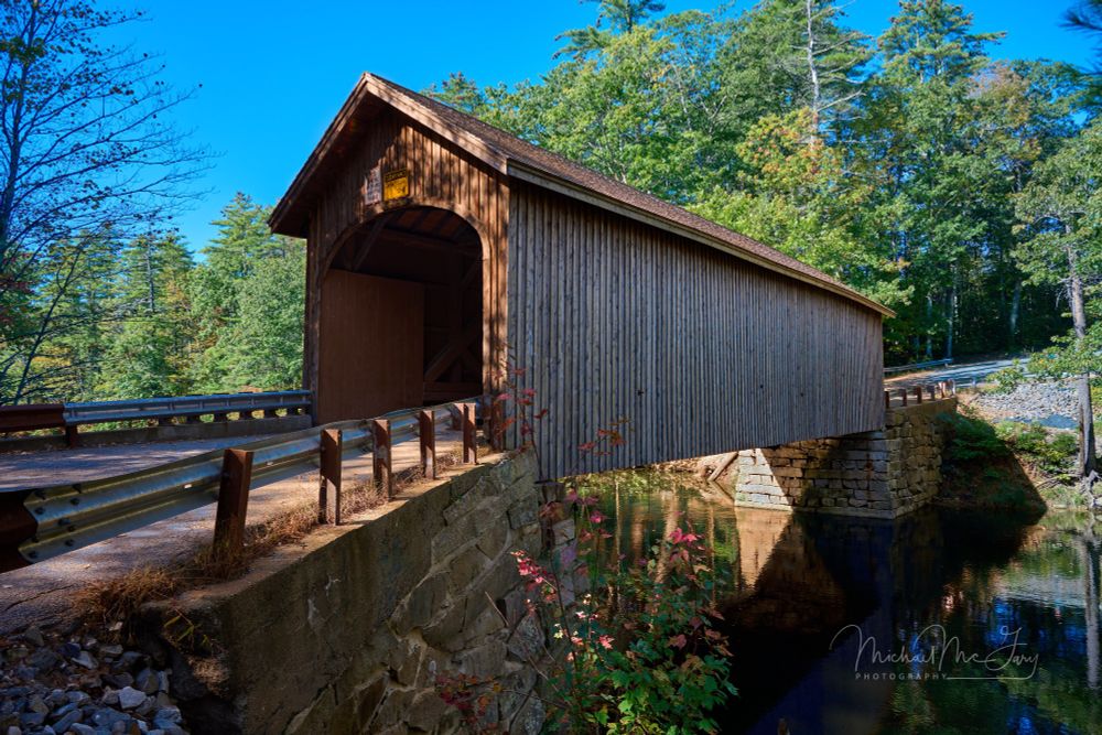 Babb's brown wood covered bridge spanning across water with green trees behind and blue sky above