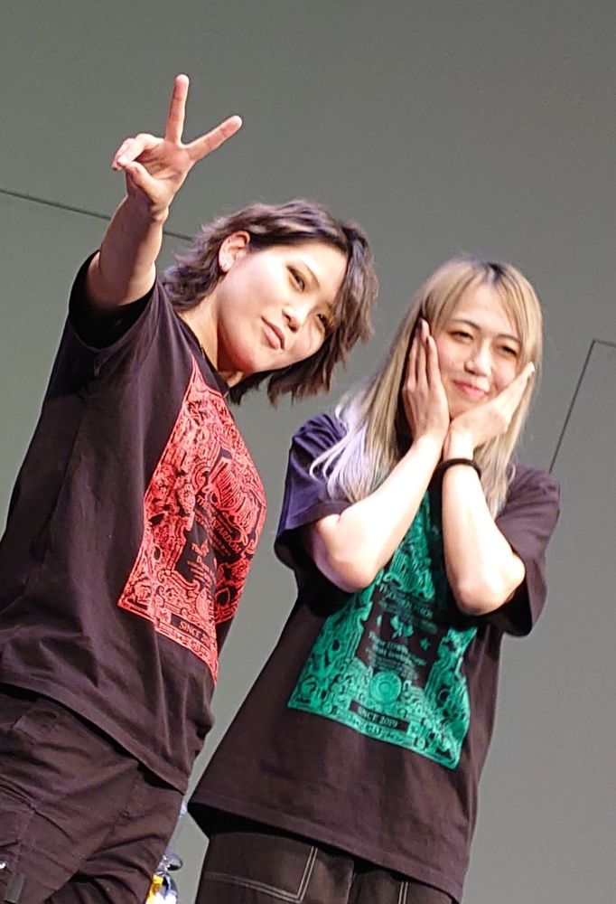 Professional wrestlers Utami Hayashishita and Saya Kamitani stand side by side, Utami cooly flashes a V with her fingers while Saya places her hands at the sides of her face in mock shock 