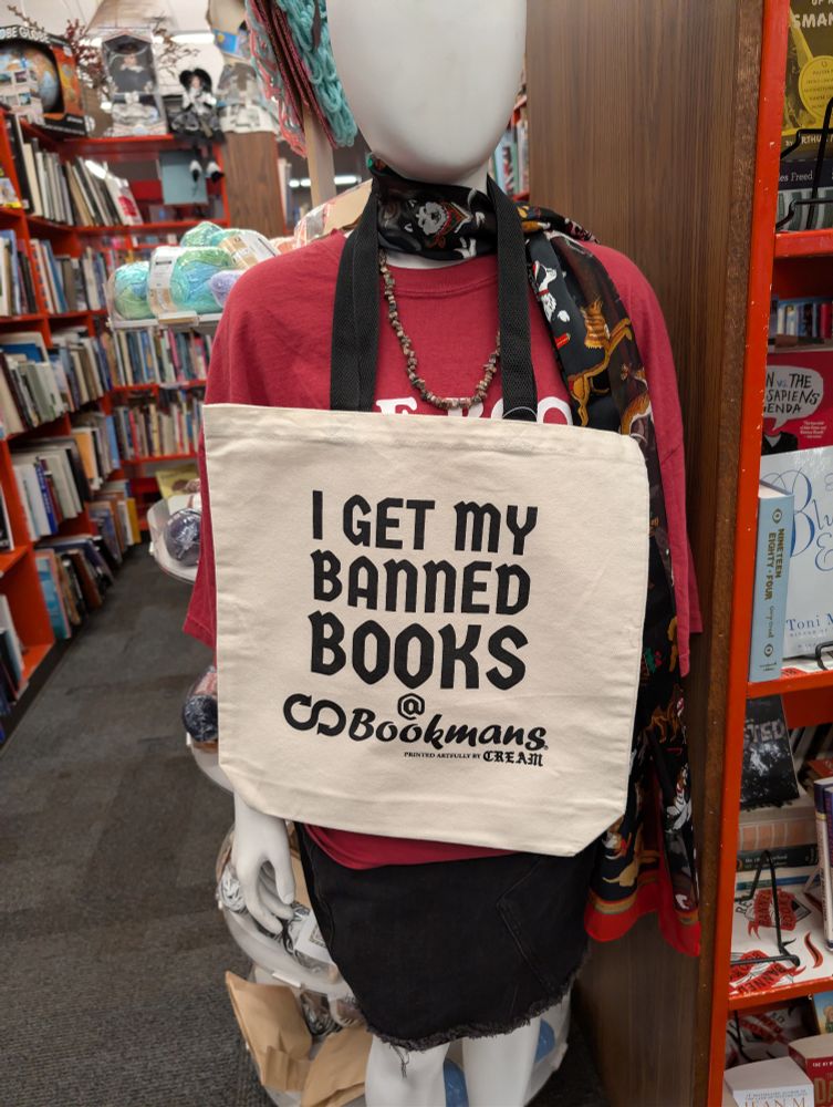 A picture I took of a mannequin with a reusable bag around its neck that says on the front "I get my banned books @ Bookmans." The bag is off white in color