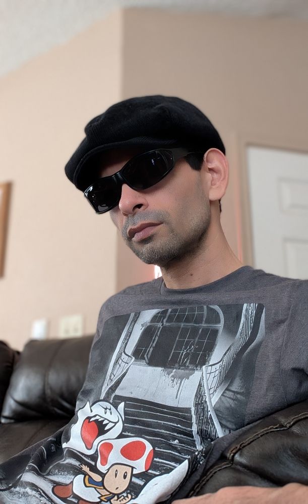 A selfie of me sitting on a couch looking serious for no reason lol. I'm wearing a black corduroy hat along with a charcoal gray t shirt with Toad from Super Mario Bros being chased by Boo ghost on the front as a print on 