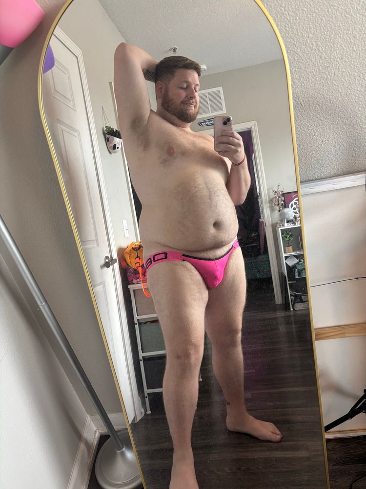 Standing in front of my mirror wearing my new neon pink low riding jockstrap with my left arm up and my hand behind my head. You can see the outline of my soft cock in the mesh material. 