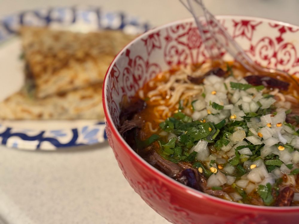 A bowl of ramenbirria soup with cilantro, onions, and peppers as toppings.