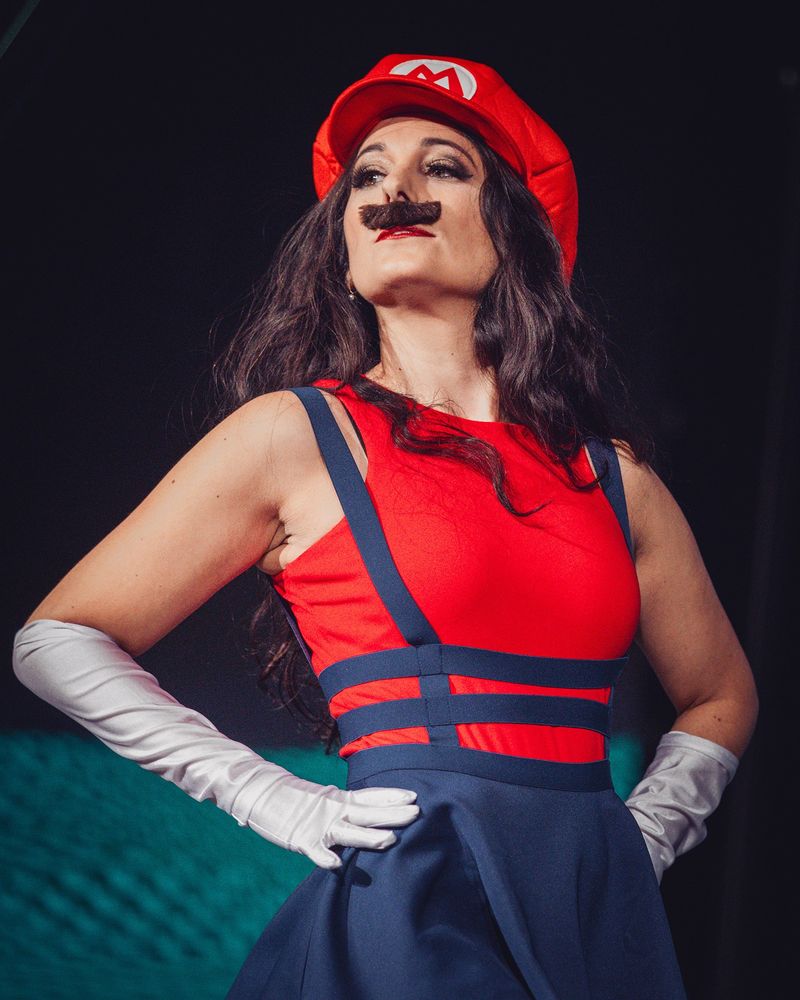 Performer "Eris Noir" stands defiantly as she cosplays Mario from Super Mario Bros. She's wearing a strappy blue skirt, red top, white gloves & signature Mario hat and mustache