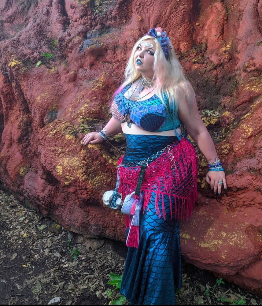 A blonde performer leaning on a rock, gazing 3/4 profile in blue mermaid garb : a mermaid-cut skirt of blue scales, a blue frilly & scaly top. Pink netting across the waist, alongside a chain belt holding a fan, mug and other knickknacks. A blue shell tiara adorns their head 