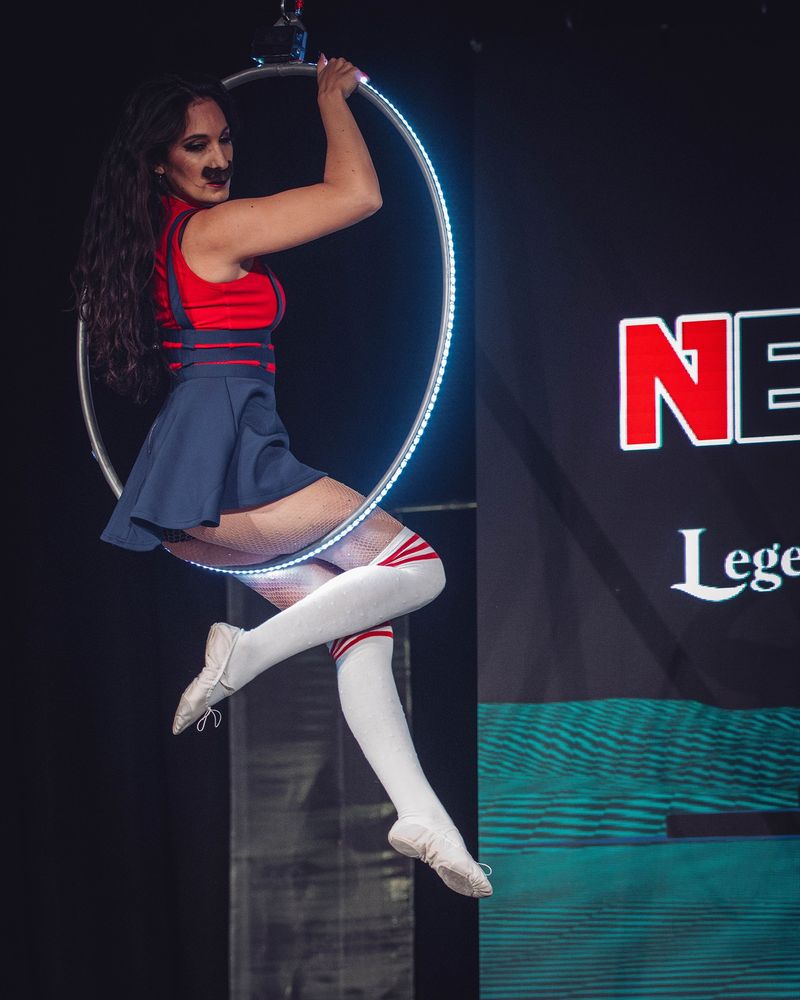 Performer "Eris Noir" on her LED Lyra apparatus. She is cosplaying Mario from Super Mario Bros. while sitting in the air on her LED hoop.
