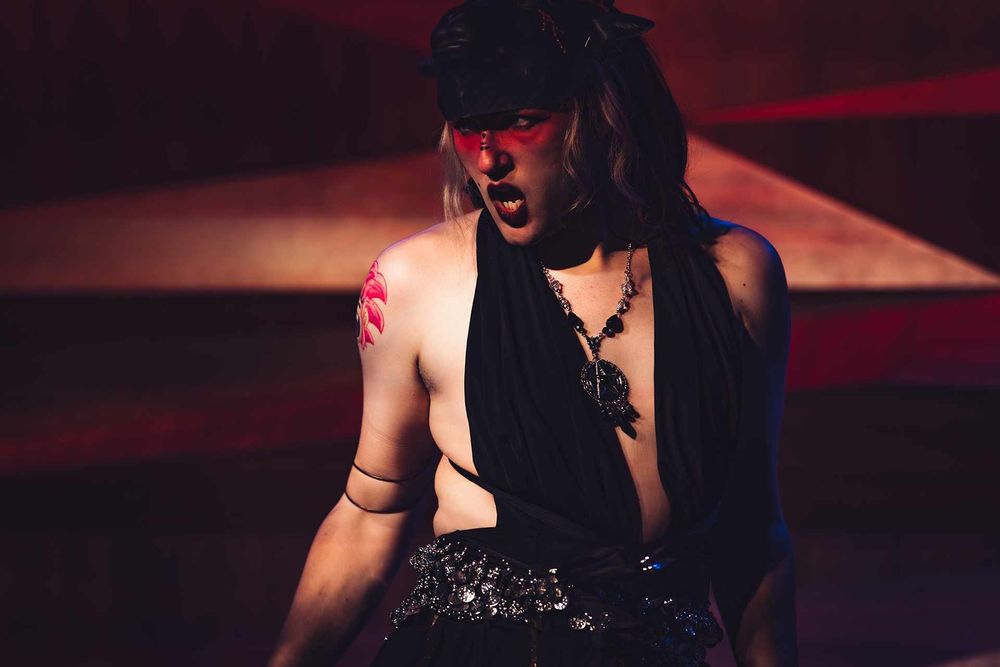 Performer "Nova Auron" dressed in a black belly dancer outfit, with red face paint & a black goat mask. They're sneering towards the left, as they are facing profile.