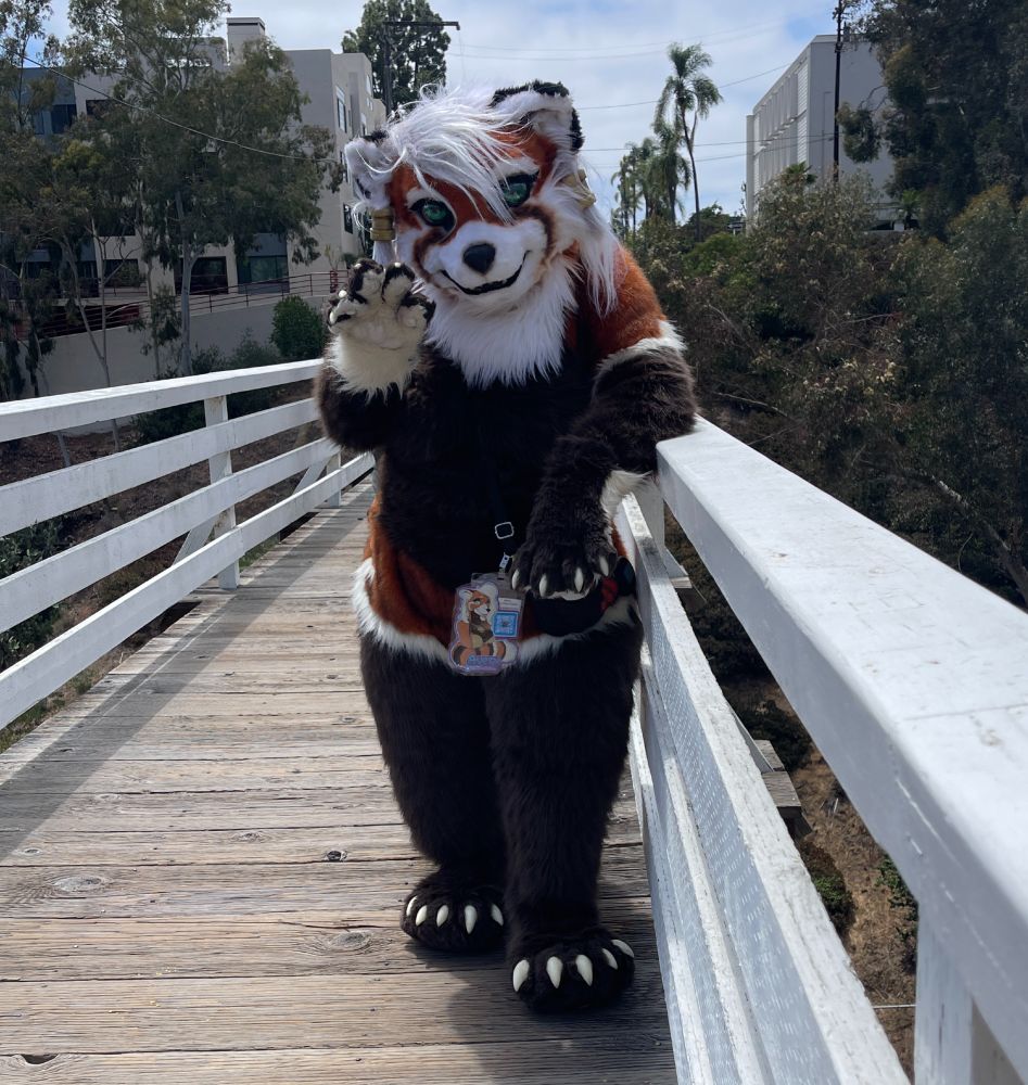 Avery the Red Panda stands on a wooden bridge that stretches across a canyon with green trees and buildings on the other side of the bridge behind her while waving a paw at the viewer.