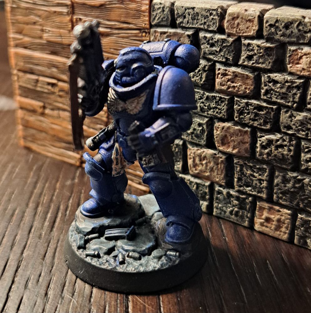 A blue space marine reloading their gun and looking to the side. They have a winged silver skull on their chest, a silver skull on the feont of their helm and black and iron accents on their belt. They are standing on a grey rocky dusted base with bullet shells and a wood and stone wall behind them.