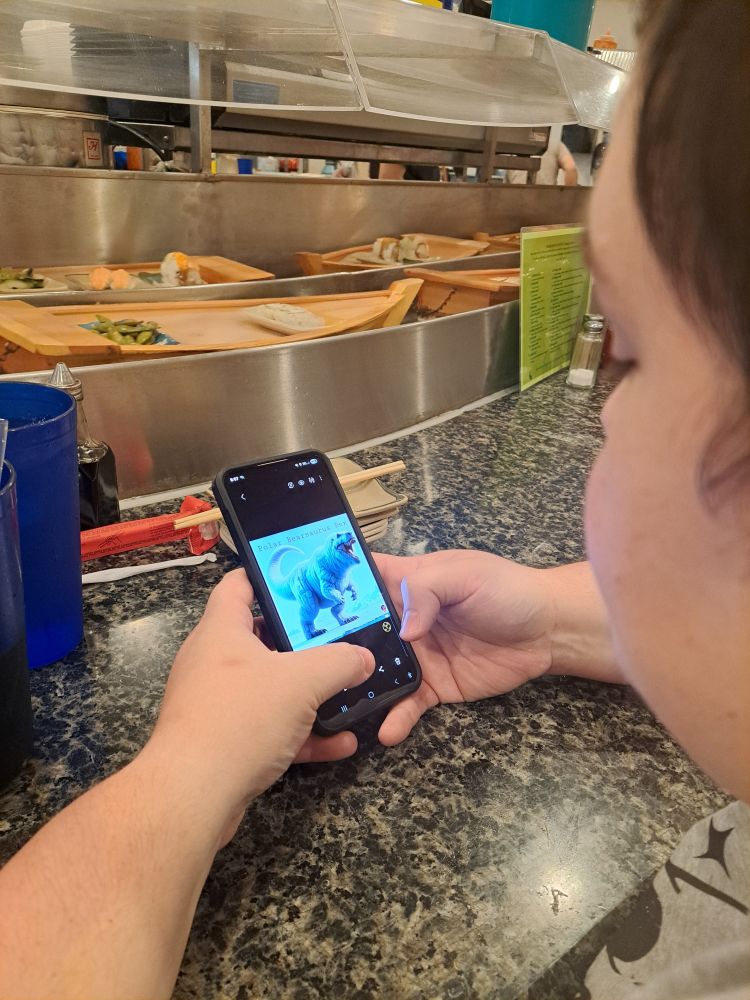 Over the shoulder picture of theo a short haired butch lesbian looking at their phone showing a polar trex. Past their phone is a conveyer belt of little boats holding various sushi.