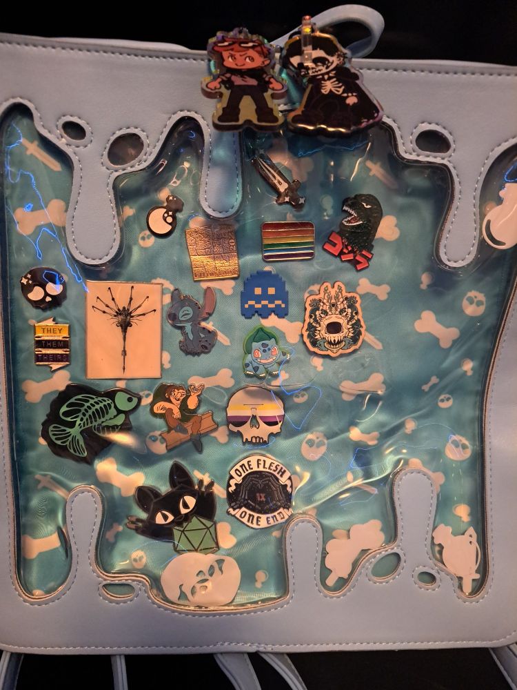 A light blue gelatinous cube ITA bag, it has variois pins in it. Including; a black cat with a dice in its mouth, a disney stitch, a disney Quasimodo pin,  a godzilla head roaring, a bulbasaur, a xray neon fish, a facehugger, a they them theirs pin, a gay gideon sunglasses skull, one flesh one end, pac man ghost, a sword, a potion, a dnd dungeon tile, and the neewest addition a wooden pin showing a skeletal beholder with mushrooms groing around it.