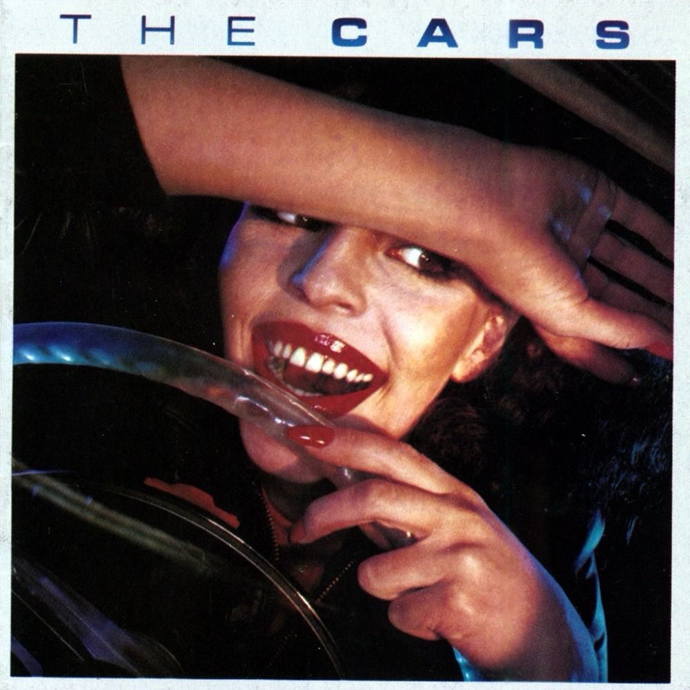 The cover of The Cars first album, showing a young, smiling woman behind the wheel of a classic custom car. 