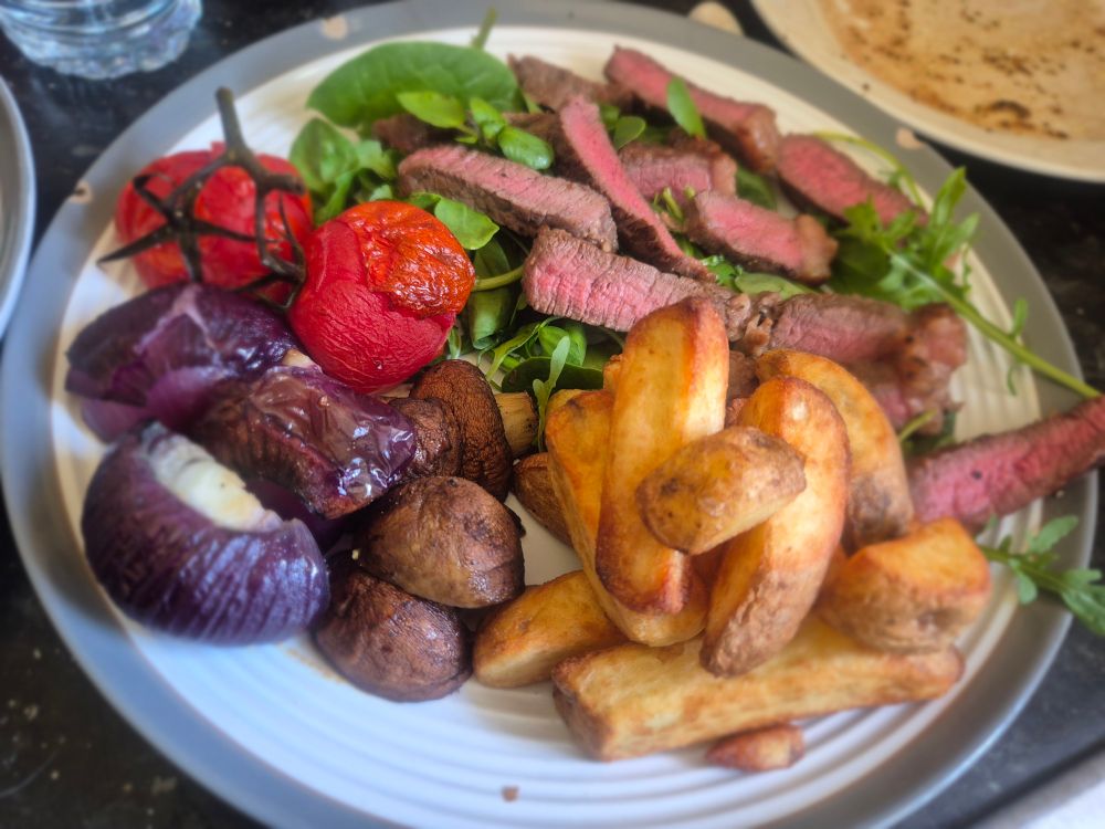 Sliced medium cooked rump steak on a bed of watercress and rocket with roasted onions, mushrooms, vine tomatoes, red onion, and a handful of chips. 