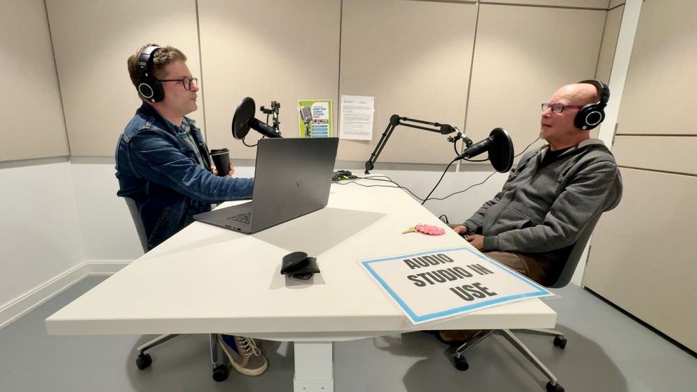 Two incredibly handsome men sit at a white table in a sound-proofed room, facing each other. They are speaking into microphones and wearing headphones.