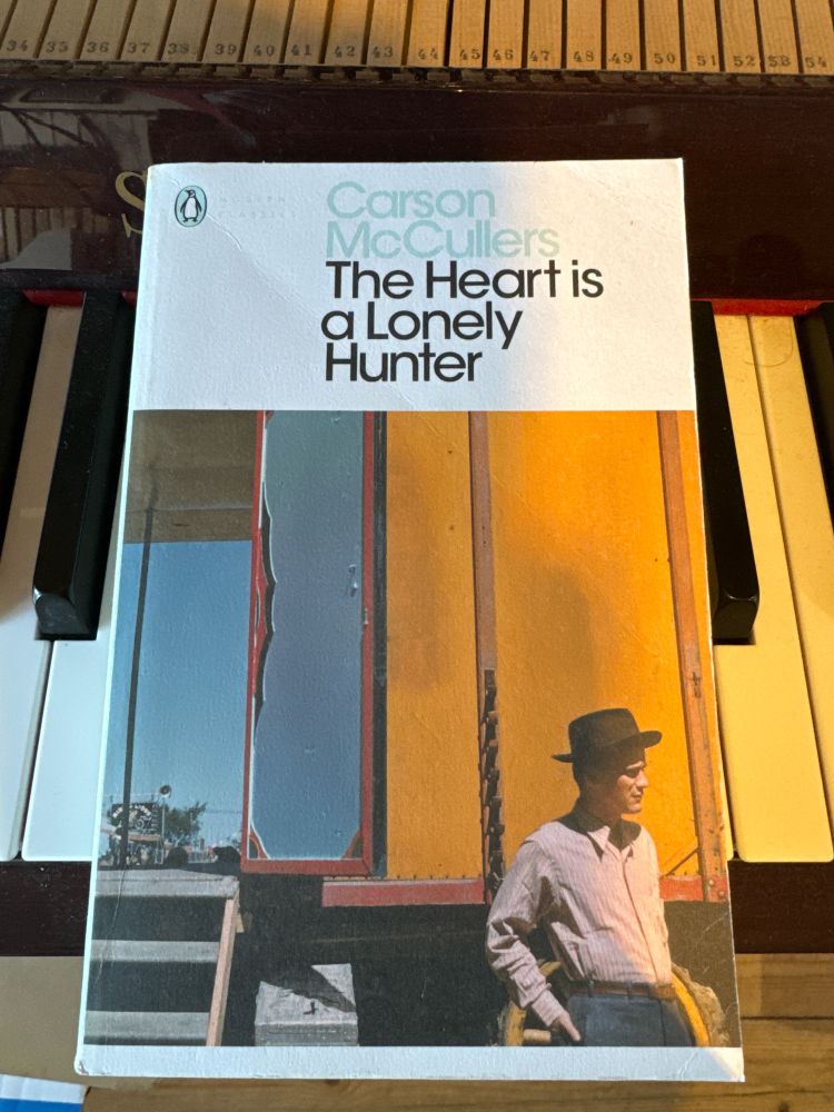 A paperback book on a piano. The book is The Heart is a Lonely Hunter by Carson McCullers and shows a cowboy type man looking away to the right with his hands in his pockets 