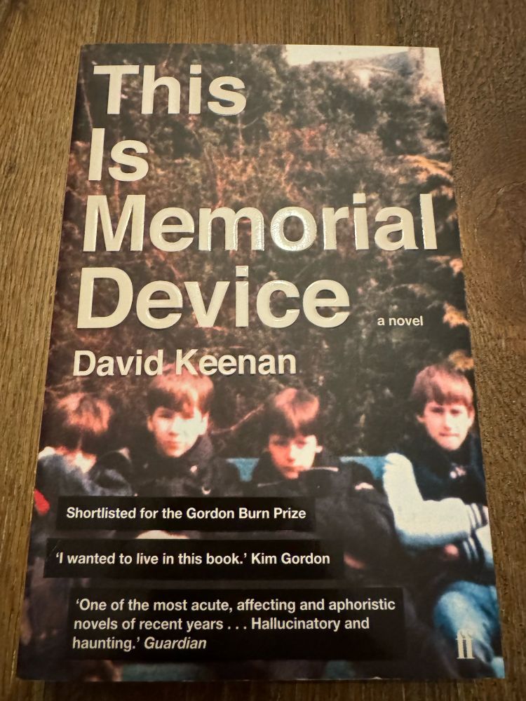 A paperback novel on a wooden floor. The novel is This Is Memorial Device by David Keenan