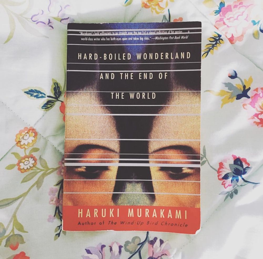 A photo of a paperback book on a floral bed spread. The book is Hardboiled Wonderland and the End of the World by Haruki Murakami and the cover depicts the corner of two faces that appear to be the mirror image of each other 