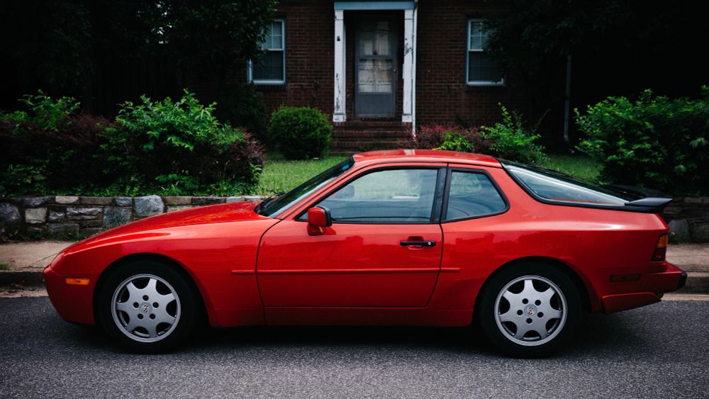 An iconic red Porsche 944 street parked in front of a brick house looking like it’s straight out of the movie “Sixteen Candles” and Jake Ryan’s about to get out.