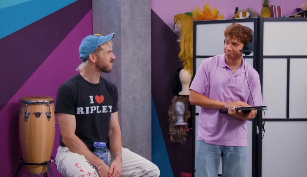 screenshot from tntl #175. tommy bowe is in the stool, and chanse mccrary is performing in a bit. tommy’s head is turned and watching chanse, who is wearing a pink shirt and black headset while holding a black leather folder. both of them are making eye contact and smiling