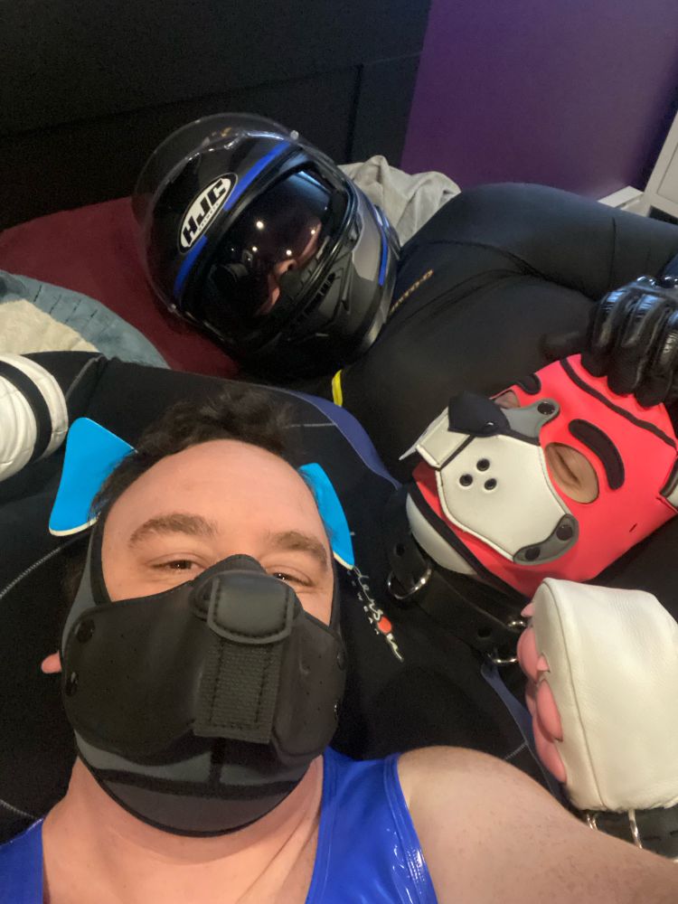 A group selfie of 3 people laying in a bed. Foreground is Plouf wearing a partial Mstr Bear puppy hood and a blue vinyl shit. Middle pup Previa is wearing a pink Mr.s hood and a wetsuit. I’m in the back and I am wearing my full face motorcycle helmet and my spandex suit. My gloved hand is laying on Previa’s head