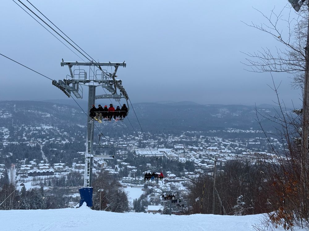 A photo showing a snowed ski run midway down the mountain. A chair lift runs through, the village and far away mountains are visible. The sky is overcast.