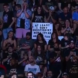 Fan holding a sign, but under herself so some words are obscured by a woman in front of her. It appears to say "at least I don't question my morality as an AEW fan"