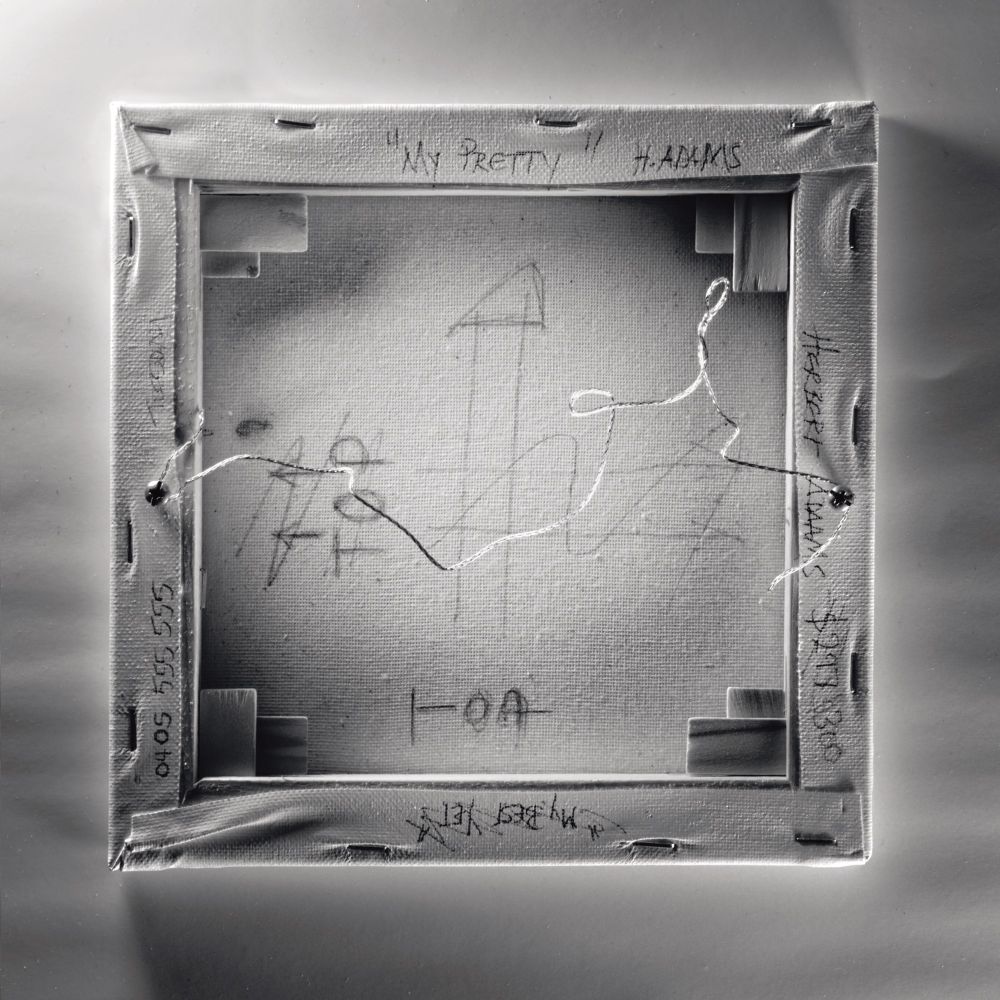 a black and white view of the rear of a small canvas that has been poorly prepared for hanging with screws central on the stretcher frame, wire twisted and way to long, nasty sharp wire ends ready to cut an errant finger, blood stains (from the curator) and various information name, phone, title scrawled on the canvas including the hanging direction(s).