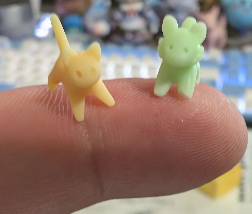 Two very small figurines, a yellow cat and a light green tiger, are sitting on the tip of an index finger.
